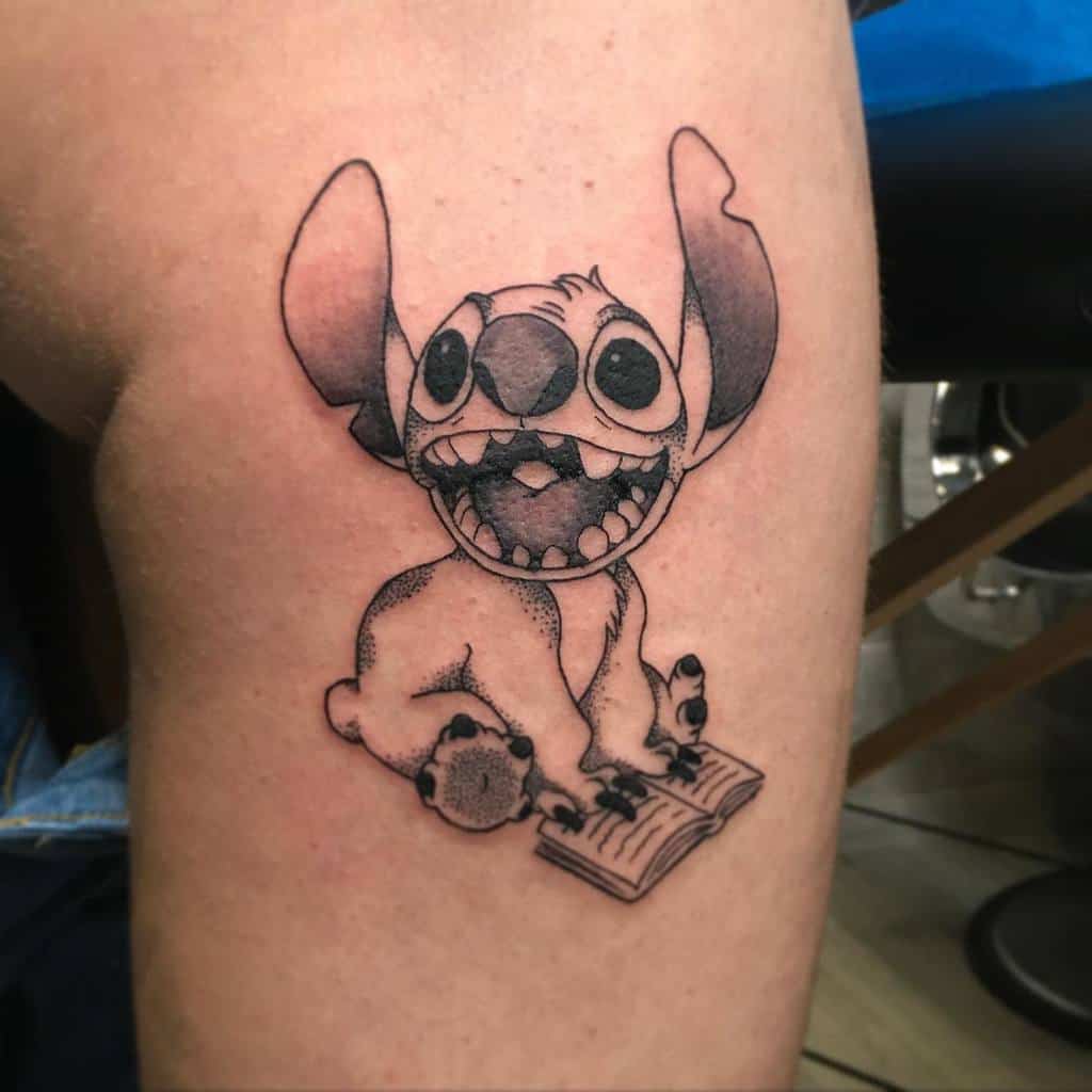 Black and white tattoo of Stitch from Lilo & Stitch, sitting on an open book with a big smile and playful expression.