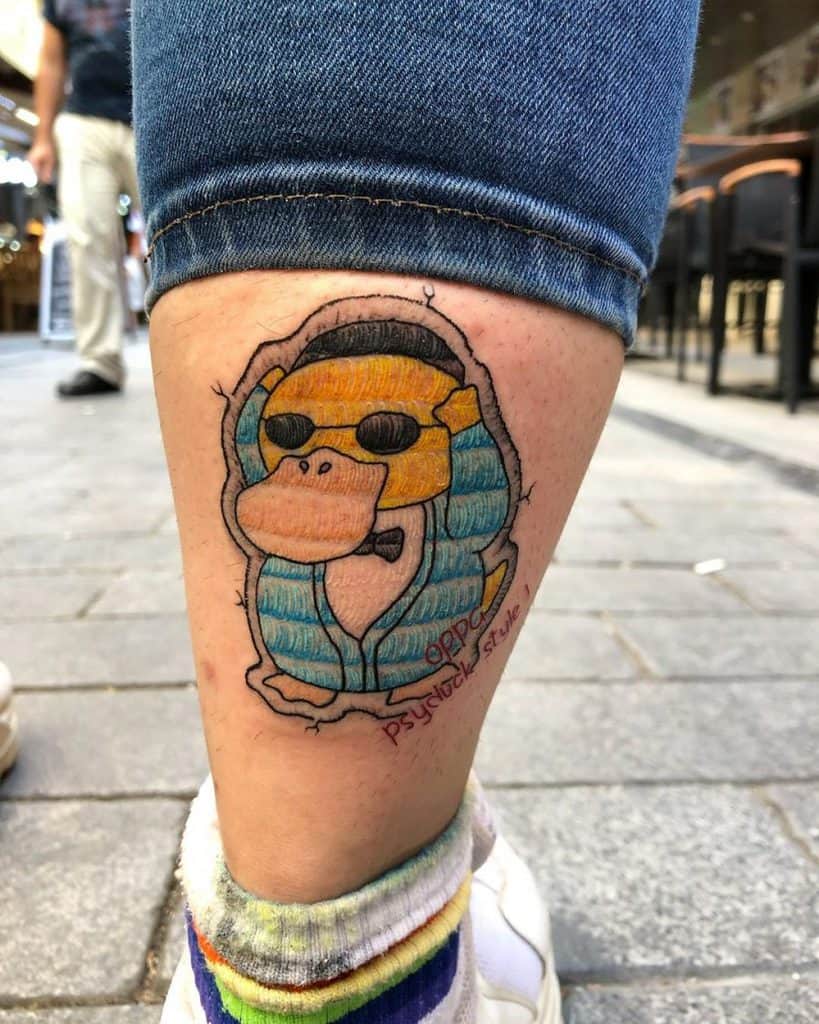 Tattoo of a Psyduck wearing sunglasses and a pharaoh headdress, designed with a fabric-like embroidery effect.