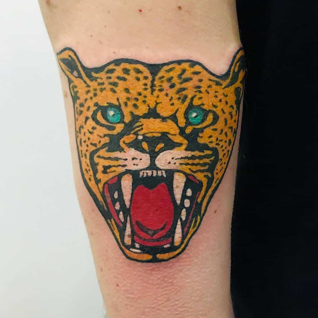 A bold, traditional tattoo of a roaring jaguar with vibrant orange and black colors, accented by striking turquoise eyes and a red mouth.
