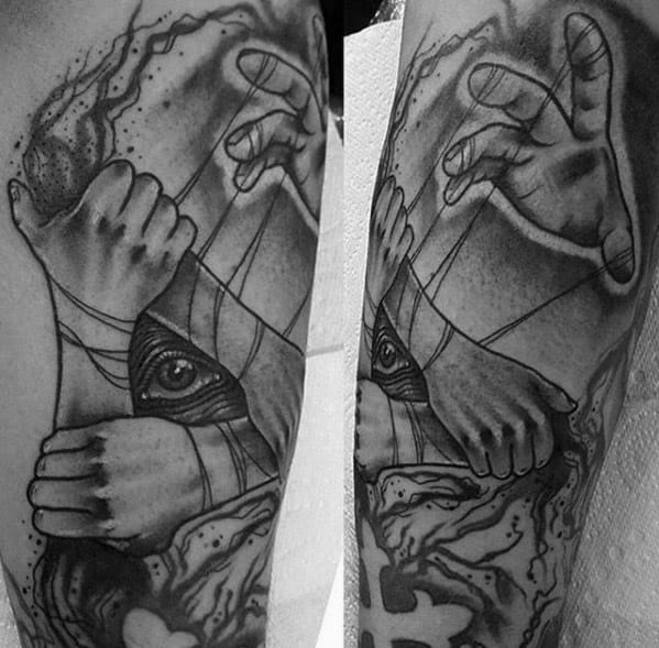 Black and grey puppet tattoo featuring hands pulling strings over a bound hand with an exposed eye, symbolizing hidden control and perception.