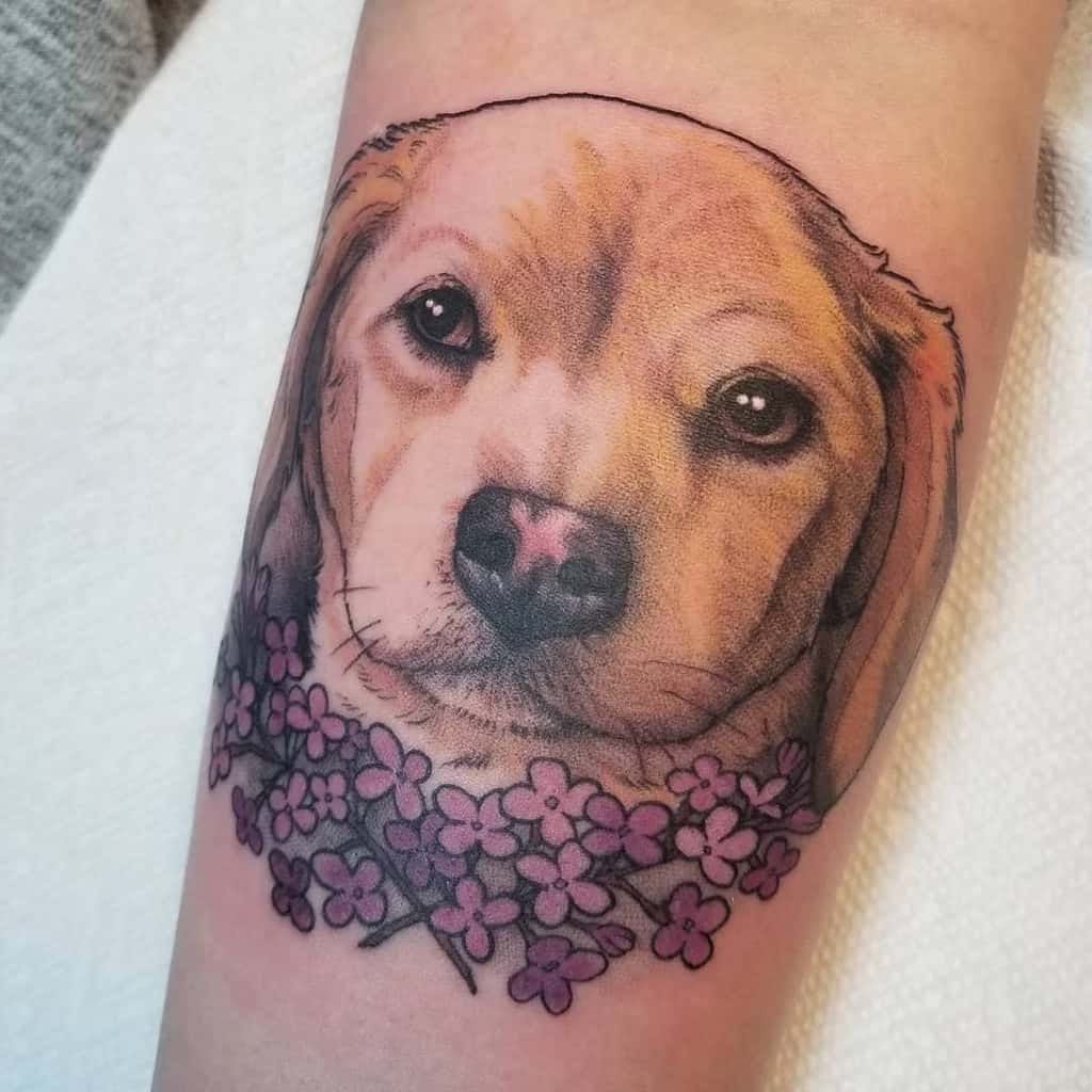 Realistic tattoo of a golden retriever puppy’s face, framed by detailed purple lilac flowers, blending soft shading and vibrant colors.