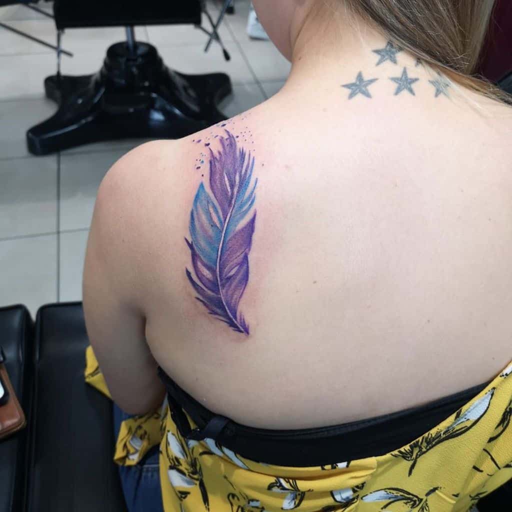 Watercolor-style feather tattoo on the upper back, featuring shades of purple and blue with soft ink splashes for an artistic effect.