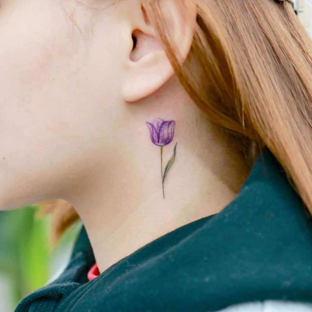 A small, delicate purple tulip tattoo behind the ear, featuring fine shading, a thin green stem, and a minimalist floral design.