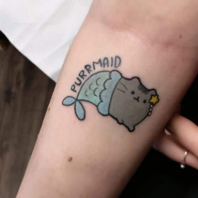 A colorful tattoo of a cute cat-mermaid hybrid with the word 'PURRMAID' above it, blending feline and aquatic themes in a playful design.