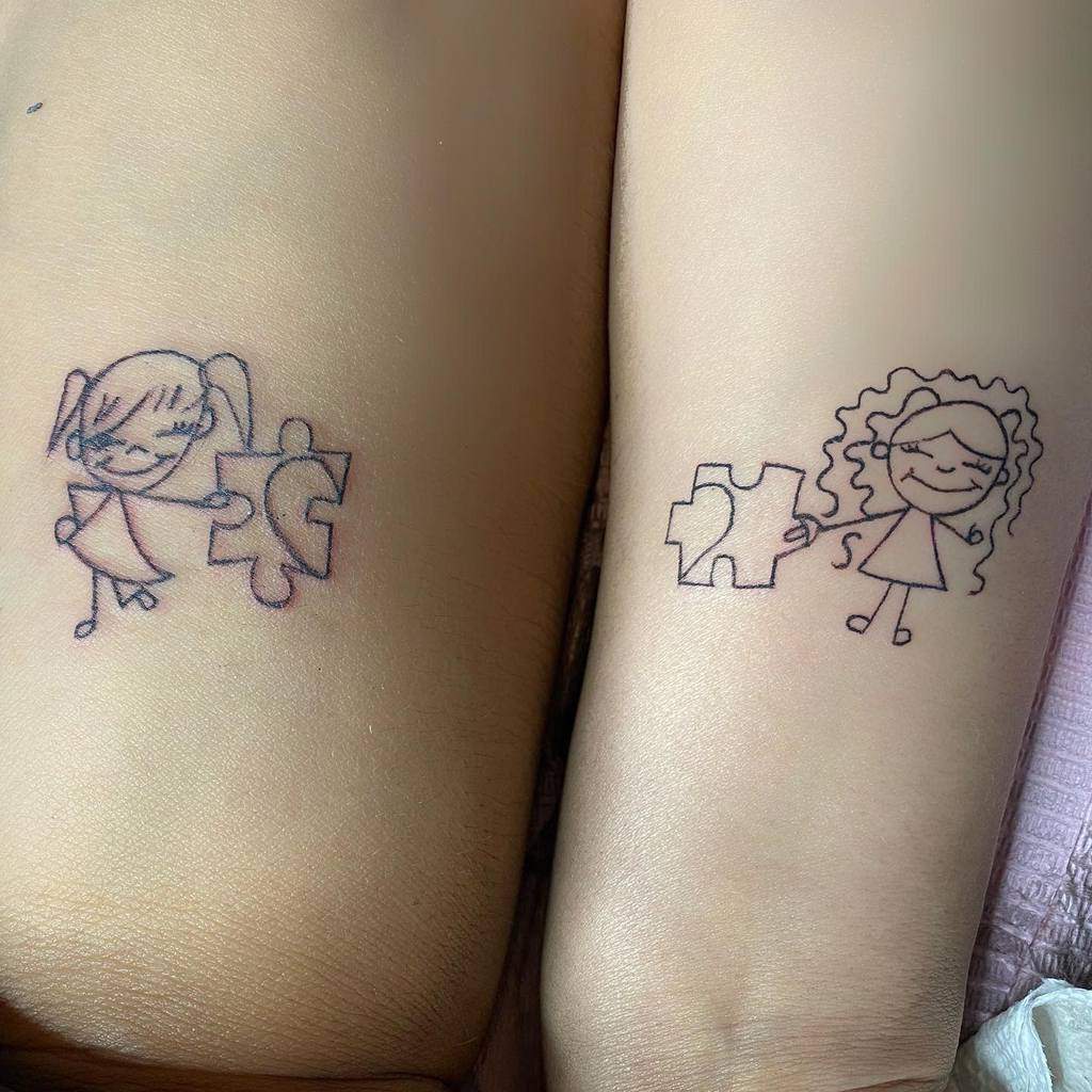 Matching sister tattoos of stick-figure girls holding puzzle heart pieces that fit together, symbolizing a strong bond and connection