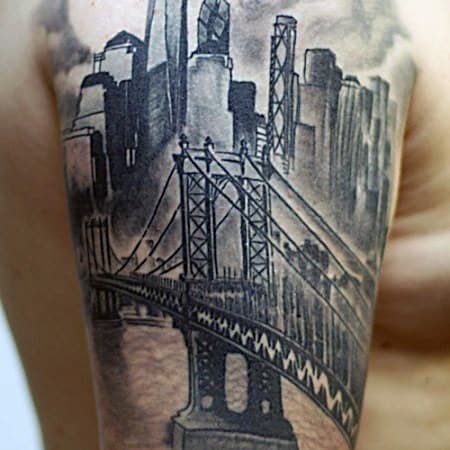 Black and gray upper arm tattoo of a New York City bridge leading into a detailed skyline with towering skyscrapers and cloudy sky.