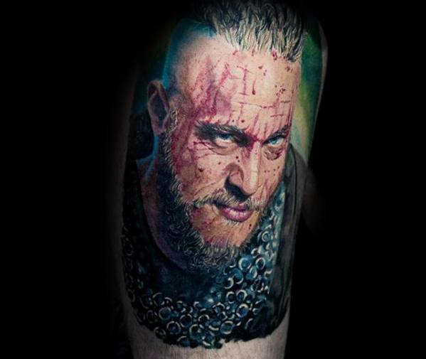 Vivid color tattoo of Ragnar Lothbrok with blood on his face and wearing chainmail armor.