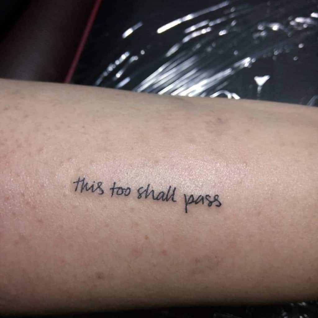 This image shows a tattoo on a person's inner forearm with the phrase "this too shall pass" in a cursive font. The tattoo appears to be freshly done with some redness around it.