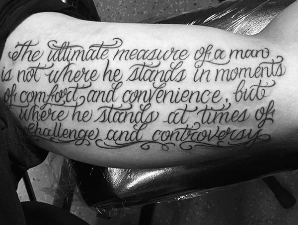 Black and grey script tattoo on the bicep featuring an elegant cursive quote with flowing, decorative lettering for a refined look.
