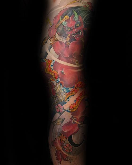 Elegant Raijin leg sleeve tattoo with red thunder god, green horns, flowing ribbons, and floral accents in a soft traditional style.