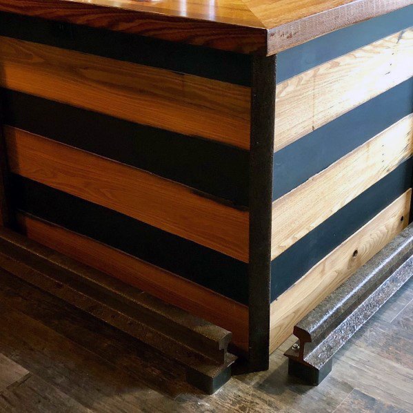 Railroad-inspired metal foot rail attached to a wooden bar with black accents on dark wood flooring.