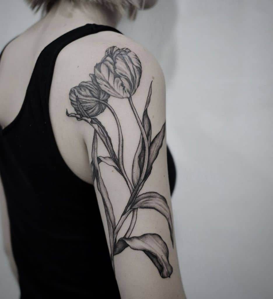 A detailed black and grey tulip tattoo covering the upper arm, featuring two large blooming flowers with intricate shading and flowing leaves.