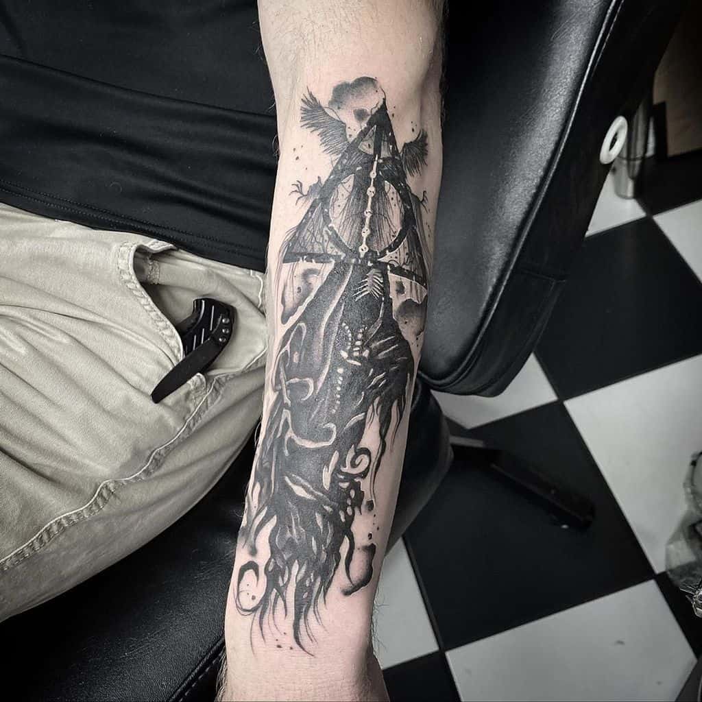 Dark-themed Deathly Hallows forearm tattoo featuring a hooded skeletal figure, skull, wings, and flowing shadows in bold black ink.