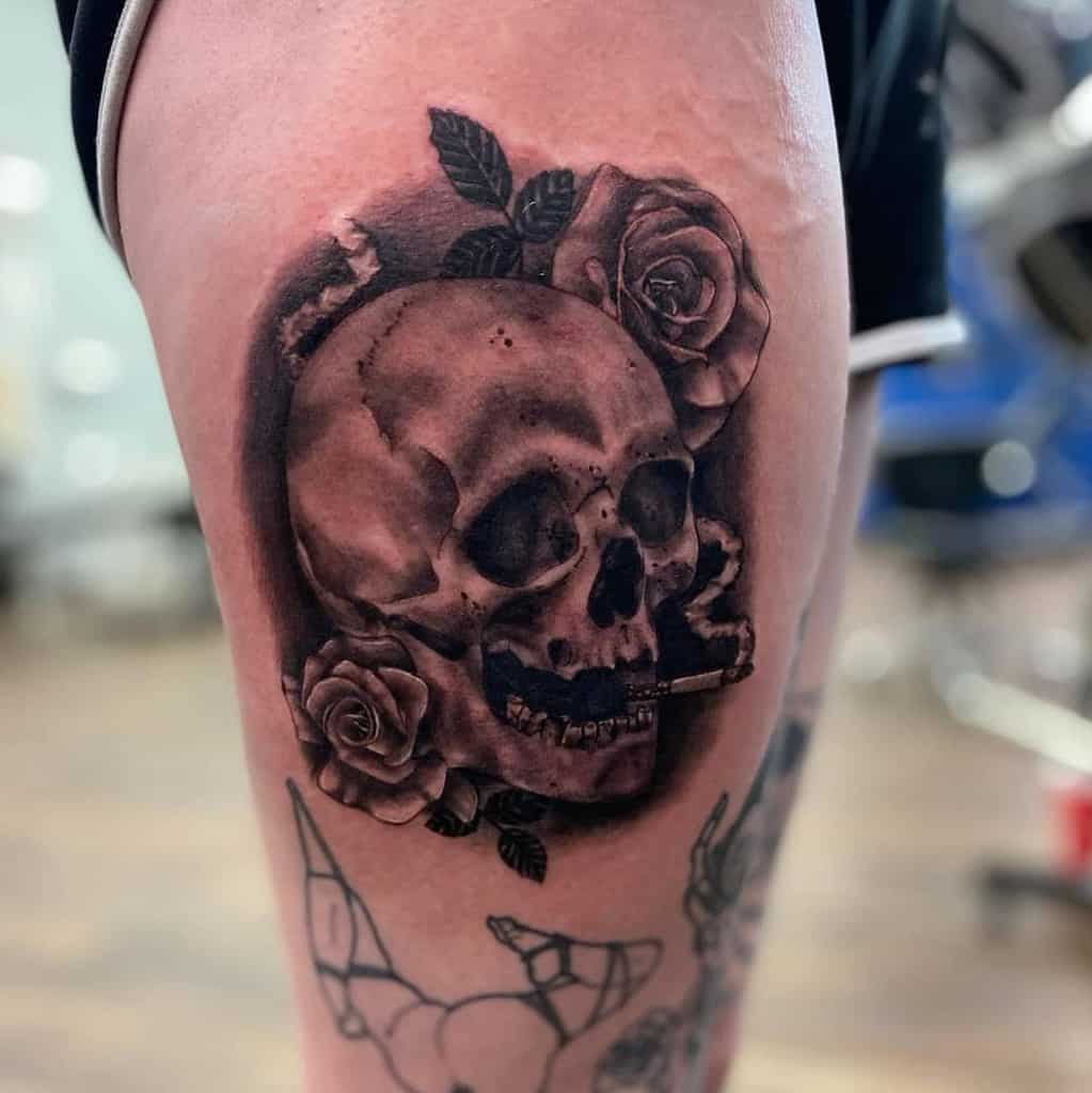 Black and grey thigh tattoo of a realistic skull smoking a cigarette, surrounded by detailed roses and deep shading for a bold look.
