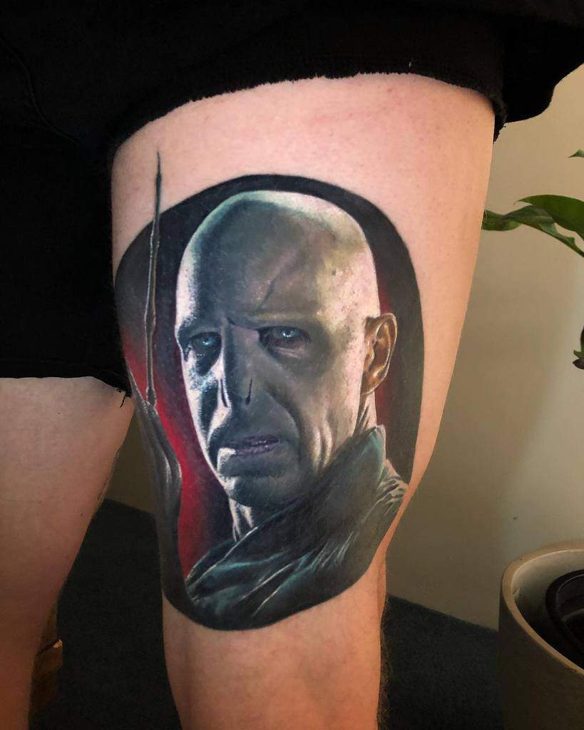 Realistic tattoo of Lord Voldemort with piercing blue eyes, holding his wand, set against a dark and red-toned background.