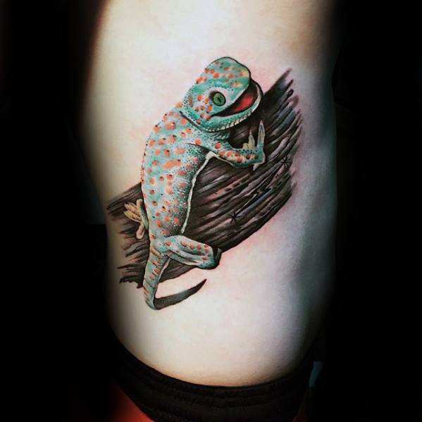 Colorful gecko tattoo with teal and orange spots, resting on a shaded wooden texture, creating a vivid and realistic look.