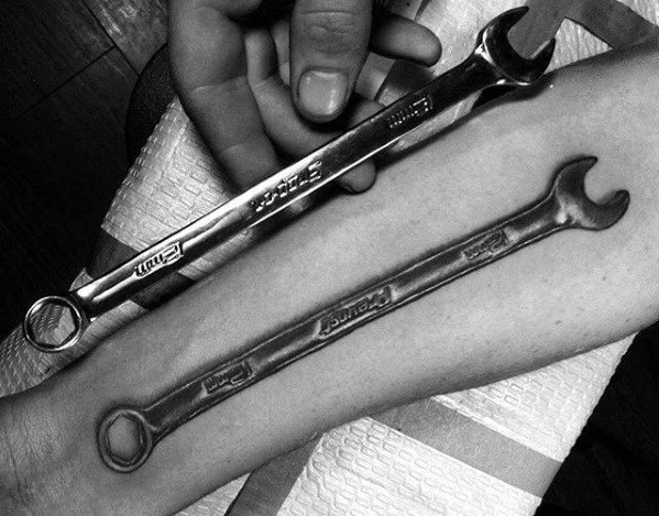 Black and grey hyper-realistic tattoo of a double-ended wrench on the forearm, closely matching the actual tool held above it.