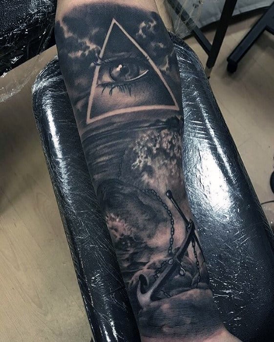 Arm tattoo featuring an eye within a triangle, waves below, and an anchor with chains in dark tones