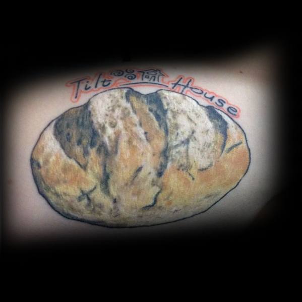 Realistic tattoo of a rustic loaf of bread with detailed shading and texture, featuring the words "Tilt House" in a bold, colorful font above it.