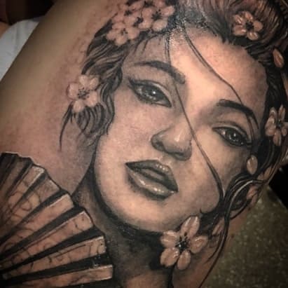 Black and gray tattoo of a Geisha with delicate facial details, flowing hair, cherry blossoms, and a partially visible hand fan.