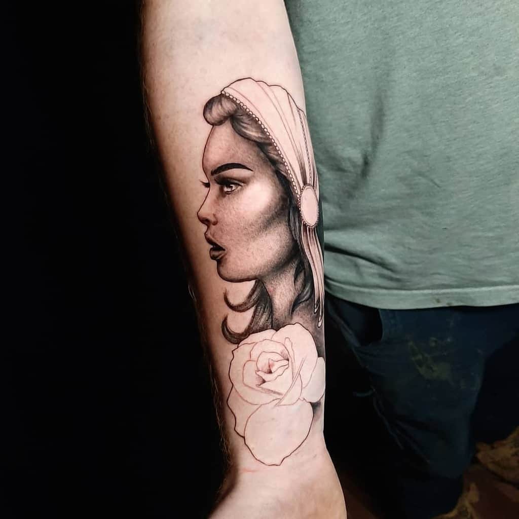 Black and grey tattoo of a gypsy woman in profile with soft shading, a headscarf, and an unfinished rose outline on the forearm.
