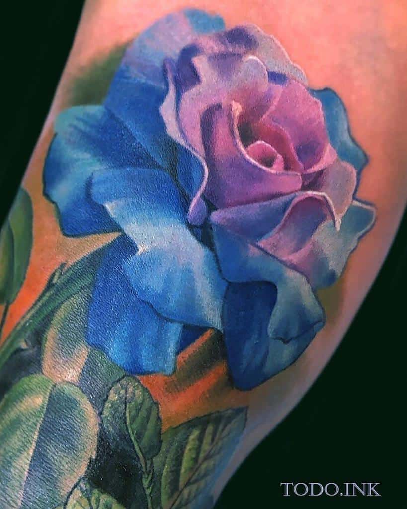 A vibrant and realistic blue and purple rose tattoo with detailed shading, placed on the arm with soft gradients and green leaves for a lifelike effect