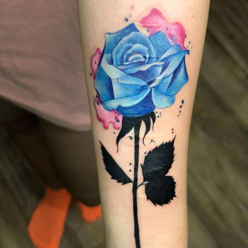 A vibrant blue rose tattoo on the forearm, with splashes of pink watercolor accents and bold black leaves, showcasing detailed shading and a dynamic style