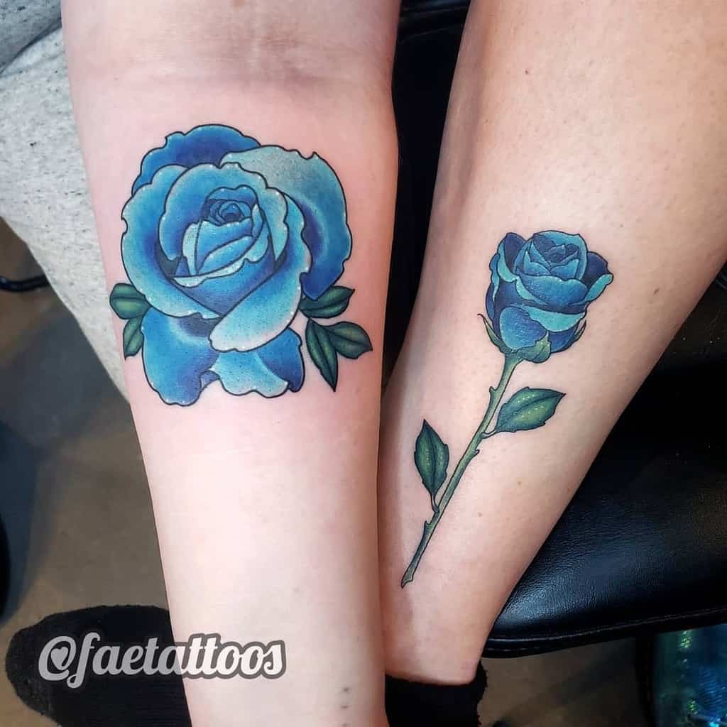 A pair of vibrant blue rose tattoos, one on the forearm with a full rose and leaves, and the other on the leg with a more minimalistic design featuring a stem