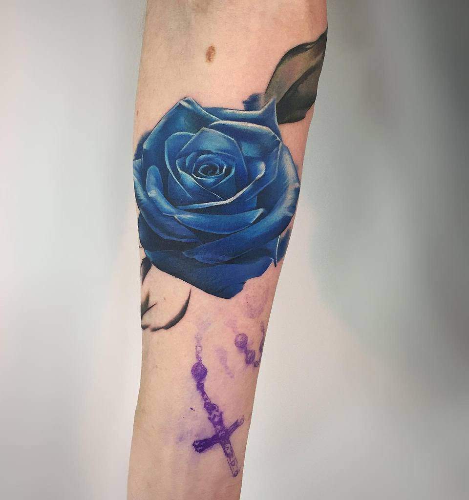 A striking tattoo featuring a vivid blue rose intertwined with a purple cross graces the person's forearm