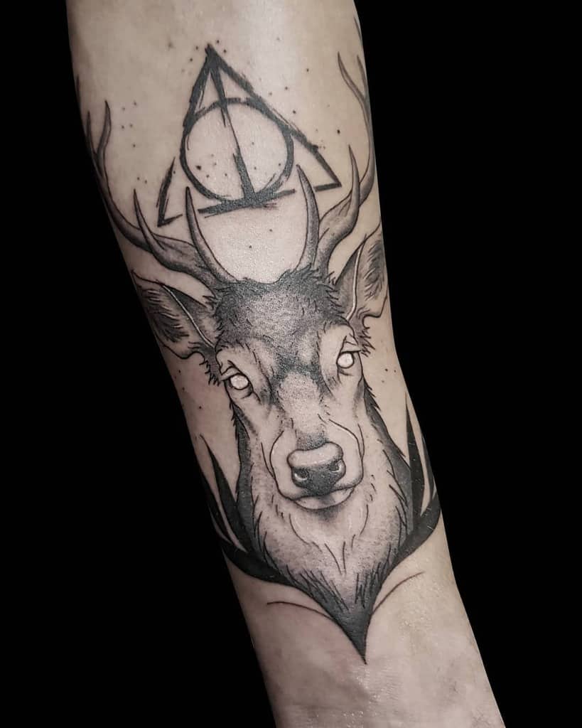 Black and grey Deathly Hallows tattoo featuring a detailed stag with glowing eyes, bold shading, and the symbol positioned above its antlers.