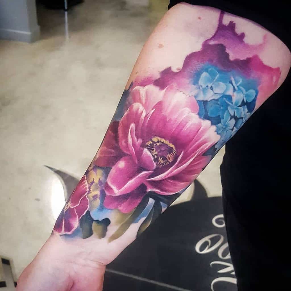 Forearm tattoo featuring vibrant pink and purple flowers, with blue accents in the background