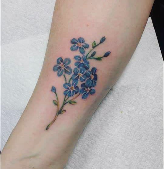A tattoo of delicate blue forget-me-not flowers with graceful green stems adorns the forearm