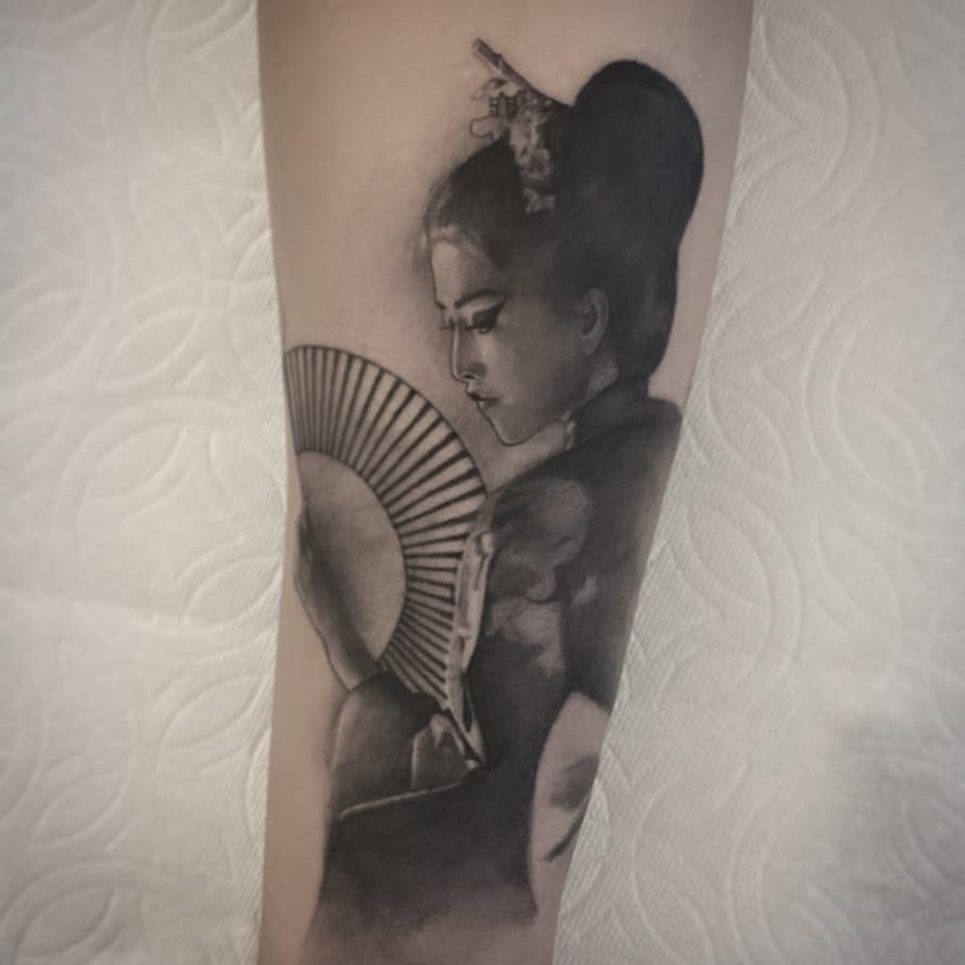 Realistic black and gray tattoo of a Geisha holding a fan, with intricate shading, a high bun, and delicate facial details.
