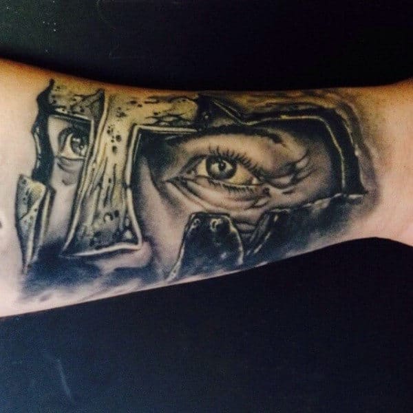 Forearm tattoo of an eye peering through a cracked surface with realistic shading, reminiscent of a gladiator's fierce gaze