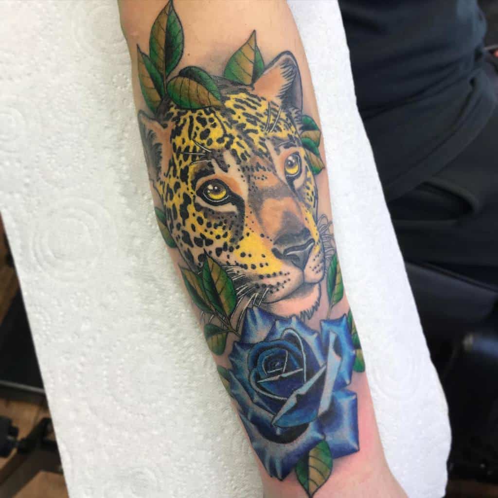 A vibrant tattoo of a jaguar’s face surrounded by lush green leaves and a bold blue rose, placed on the forearm for a colorful and detailed design.