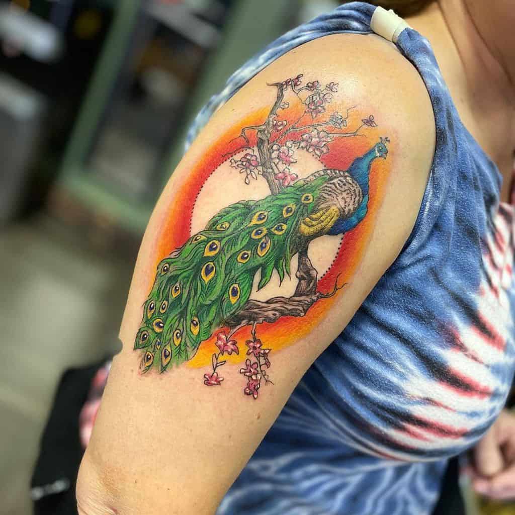 Vibrant peacock tattoo on the upper arm, with colorful feathers and a blooming tree in the background, set against a circular gradient.