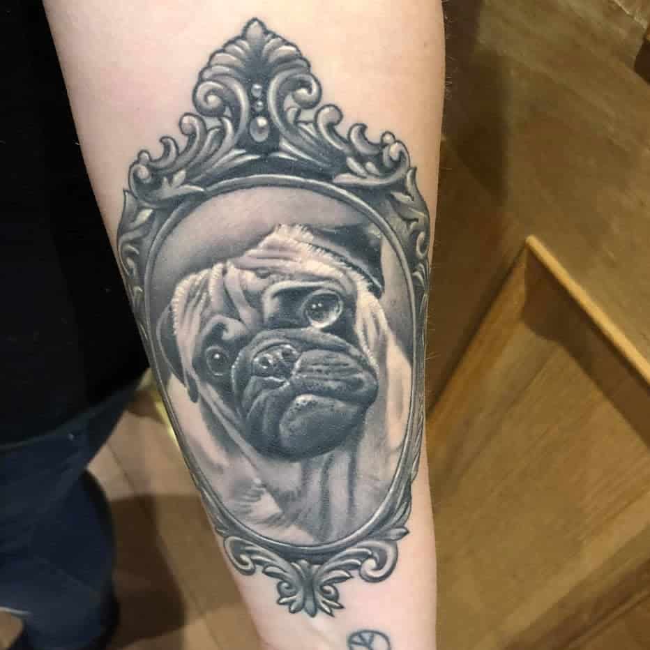 A detailed black-and-gray tattoo of a pug's face framed within an ornate, vintage-style frame