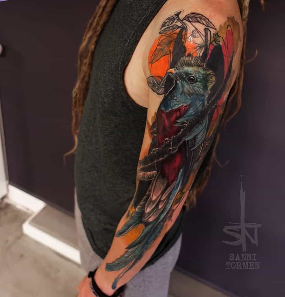 Realistic full-sleeve quetzal tattoo on the arm, featuring a vibrant green and red bird perched on a branch with intricate details and an orange background
