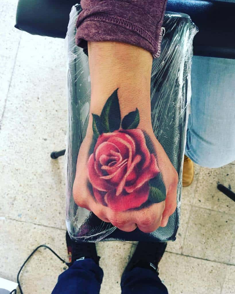 A hand tattoo showcasing a vibrant pink rose, complete with lush green leaves