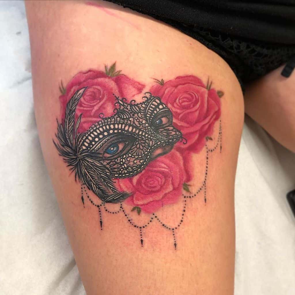 Thigh tattoo featuring a detailed lace masquerade mask with blue eyes, surrounded by vibrant pink roses and delicate beaded chains.
