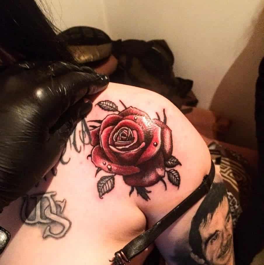 A skilled tattoo artist meticulously works on a vibrant red rose, expertly adorning the individual's shoulder with intricate detail and bold color