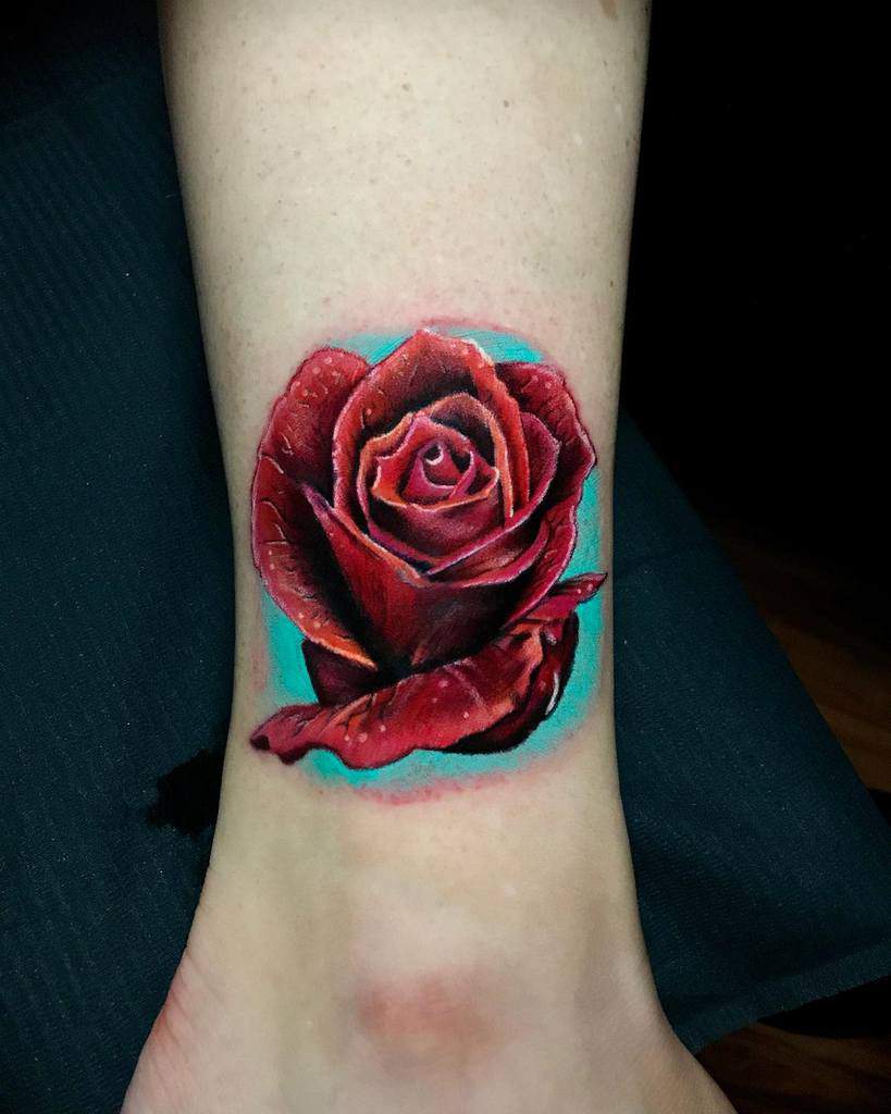 A realistic tattoo of a red rose with a teal background on an ankle