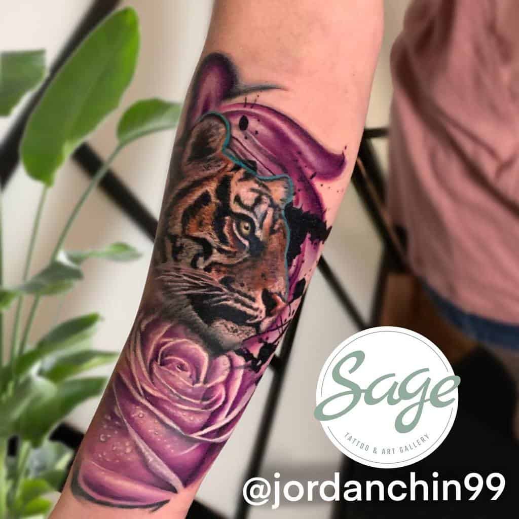 A striking forearm tattoo featuring a majestic tiger intertwined with a vibrant purple rose, proudly displaying the "Sage Tattoo & Art Gallery" logo