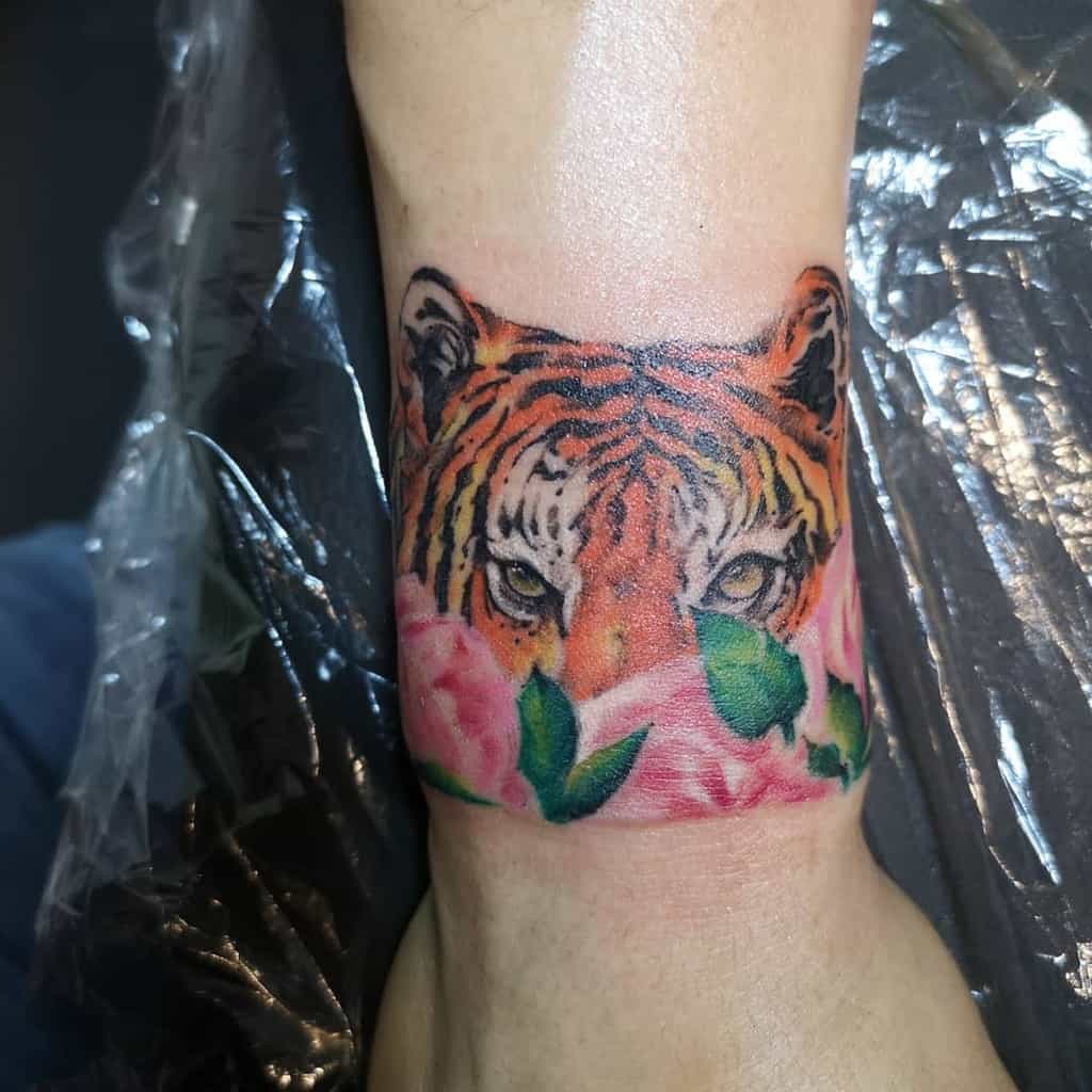 A tiger tattoo, featuring lush green leaves and vibrant pink flowers, elegantly decorates the forearm