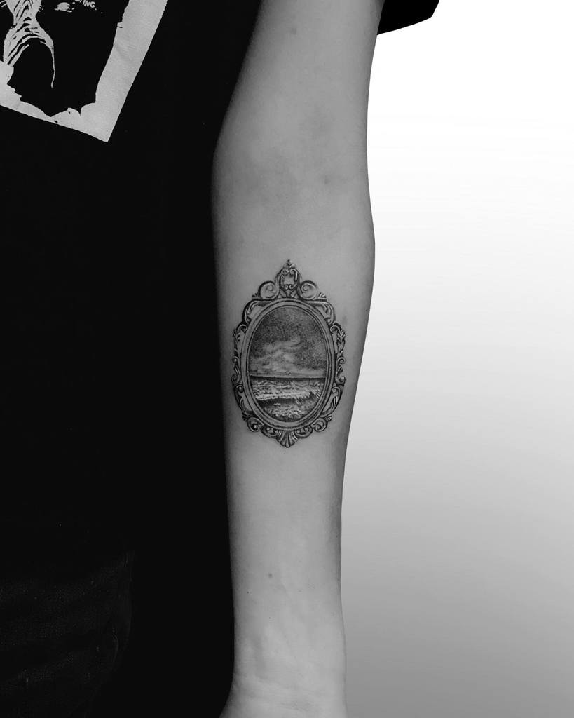Intricate tattoo featuring an ornate vintage mirror frame reflecting a serene ocean scene with waves and clouds