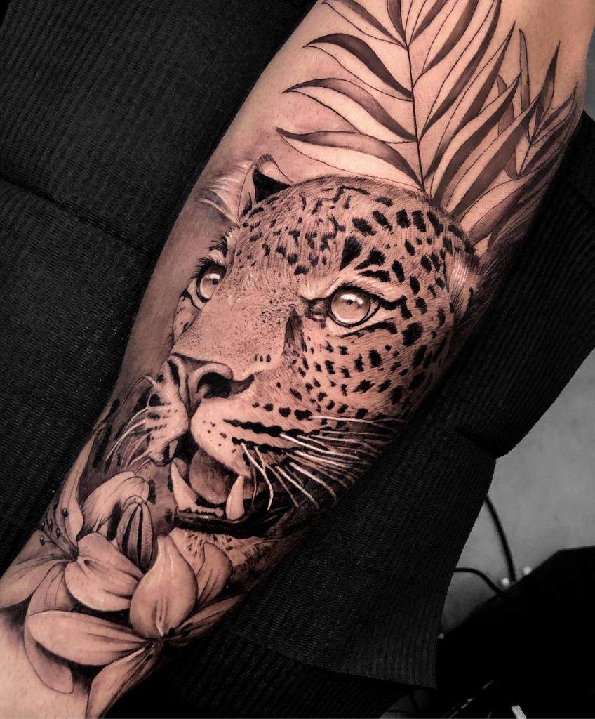 A stunning black-and-gray tattoo of a jaguar's face surrounded by lush foliage and delicate flowers, capturing both strength and beauty in intricate detail.