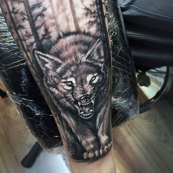 A detailed gray wolf tattoo, fierce with bared teeth, emerges from a shadowed forest backdrop on a leg
