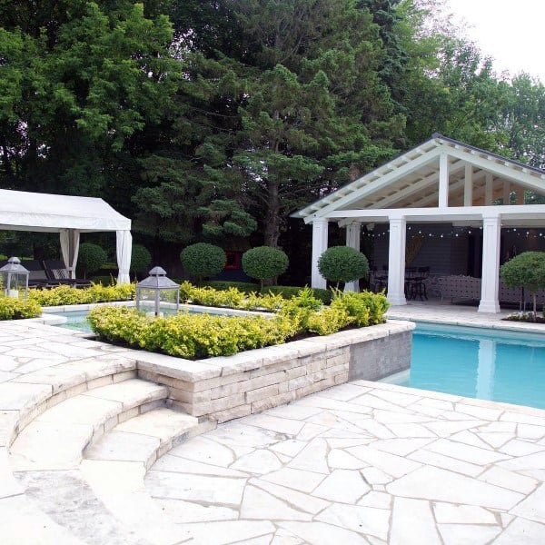 Backyard with a pool, white pergola, stone patio, lush greenery, and a shaded lounge area.