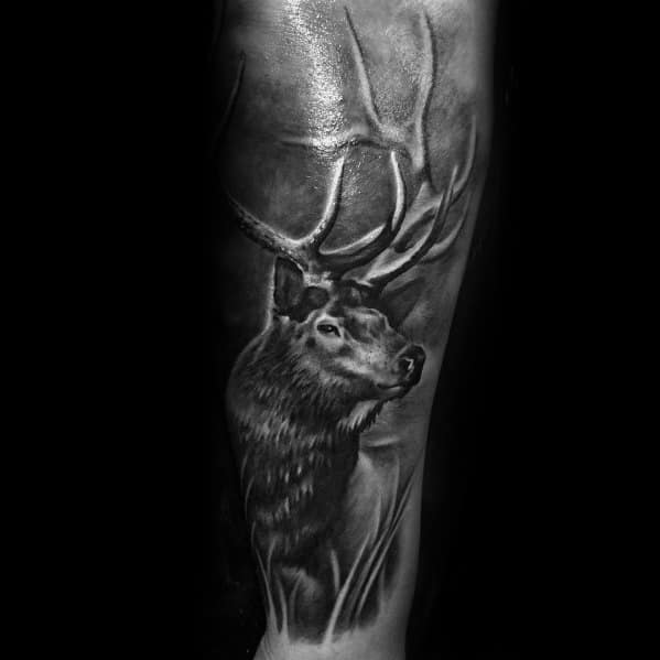 Realistic black and grey elk tattoo on the forearm, featuring intricate shading, bold antlers, and a misty background for a striking nature-inspired design.