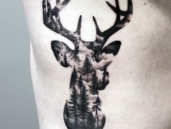 Tattoo of a deer's head silhouette filled with a forest and cloudy sky scene on skin